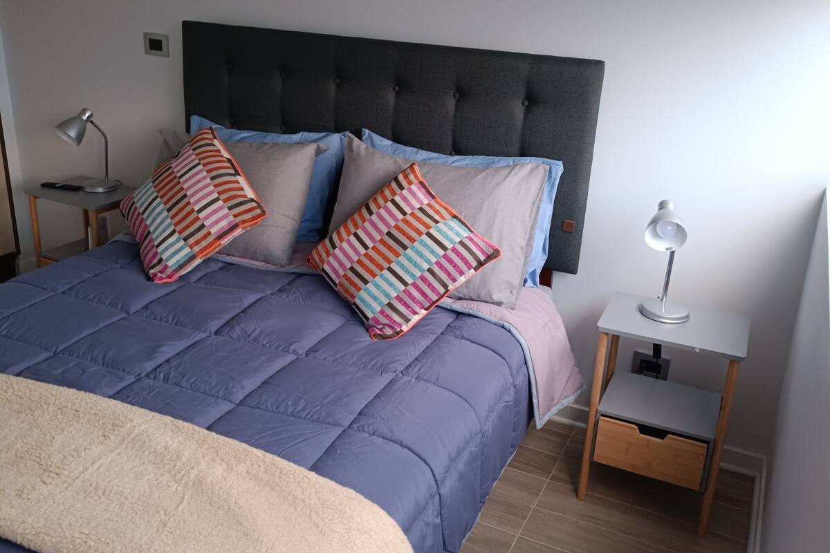 Een net opgemaakt bed met een blauwe quilt, een donker hoofdbord en gestreepte kussens. Aan beide zijden van het bed staan nachtkastjes met lampen.