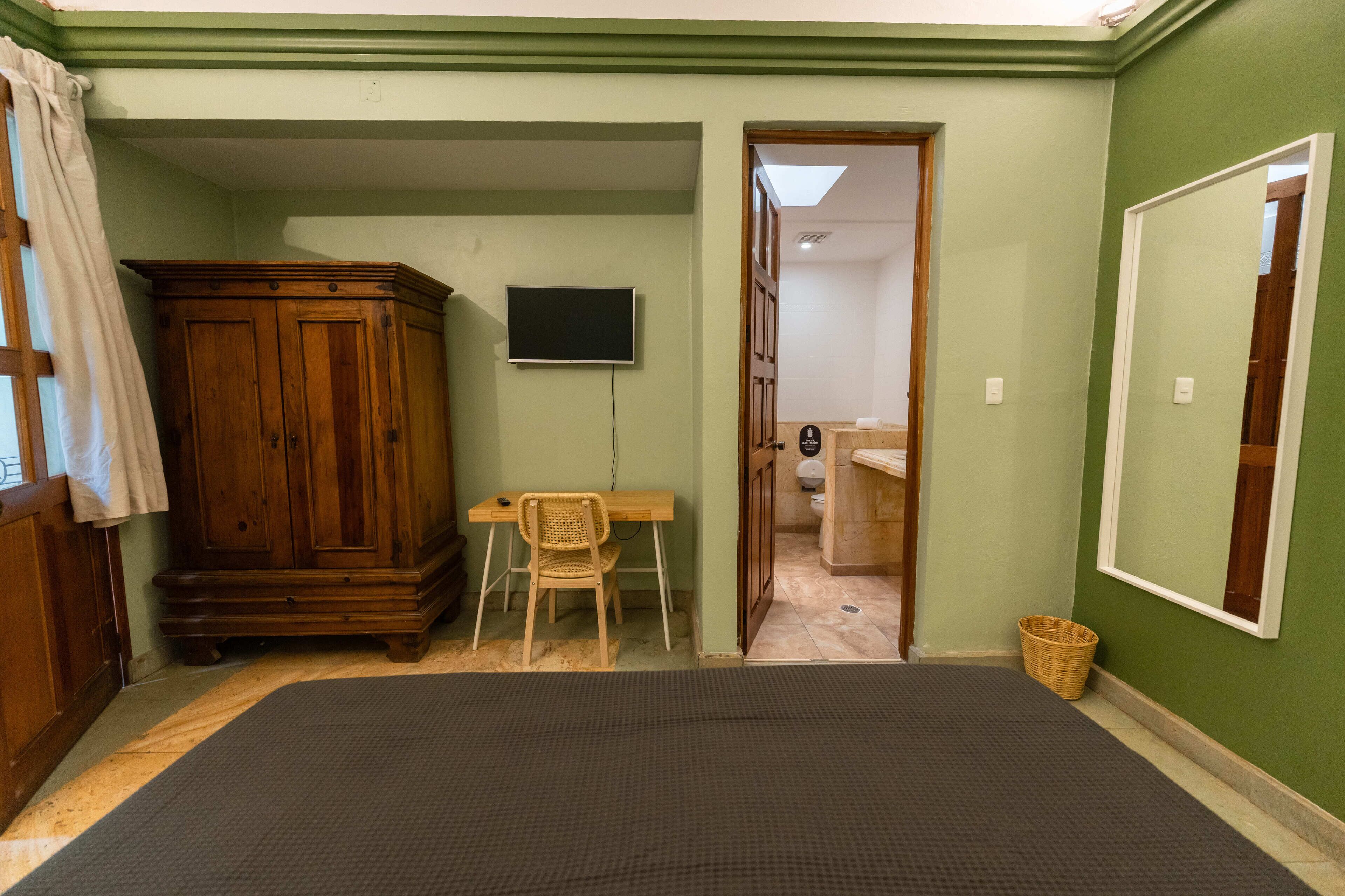 Une pièce comprenant un lit, une armoire en bois, un bureau avec une chaise, un téléviseur et une salle de bain visible à travers une porte ouverte.