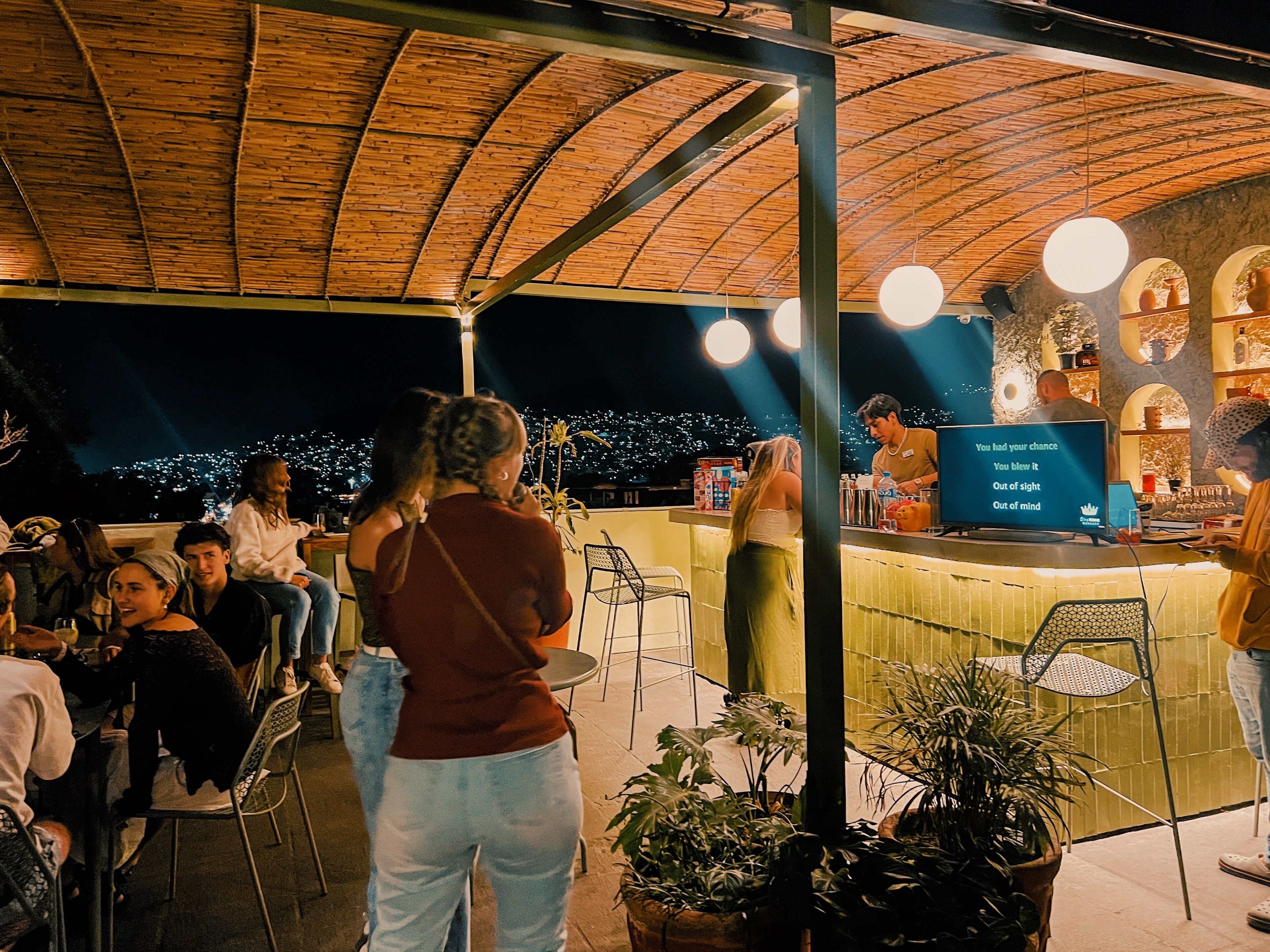 An outdoor bar area with a thatched roof, seating arrangements, and a bartender at the counter.