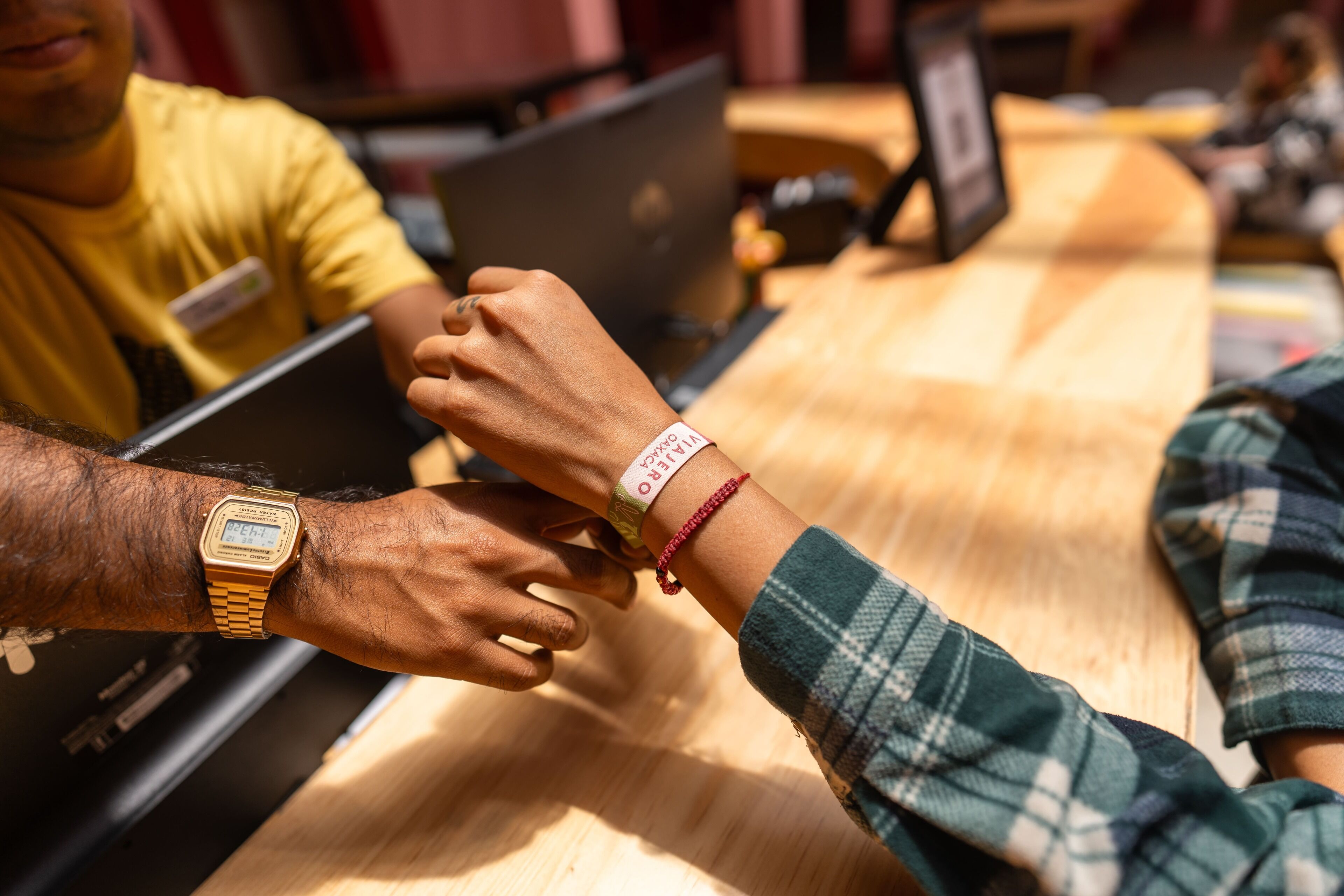 Two people sitting at a wooden table with laptops, one wearing a watch and a bracelet.