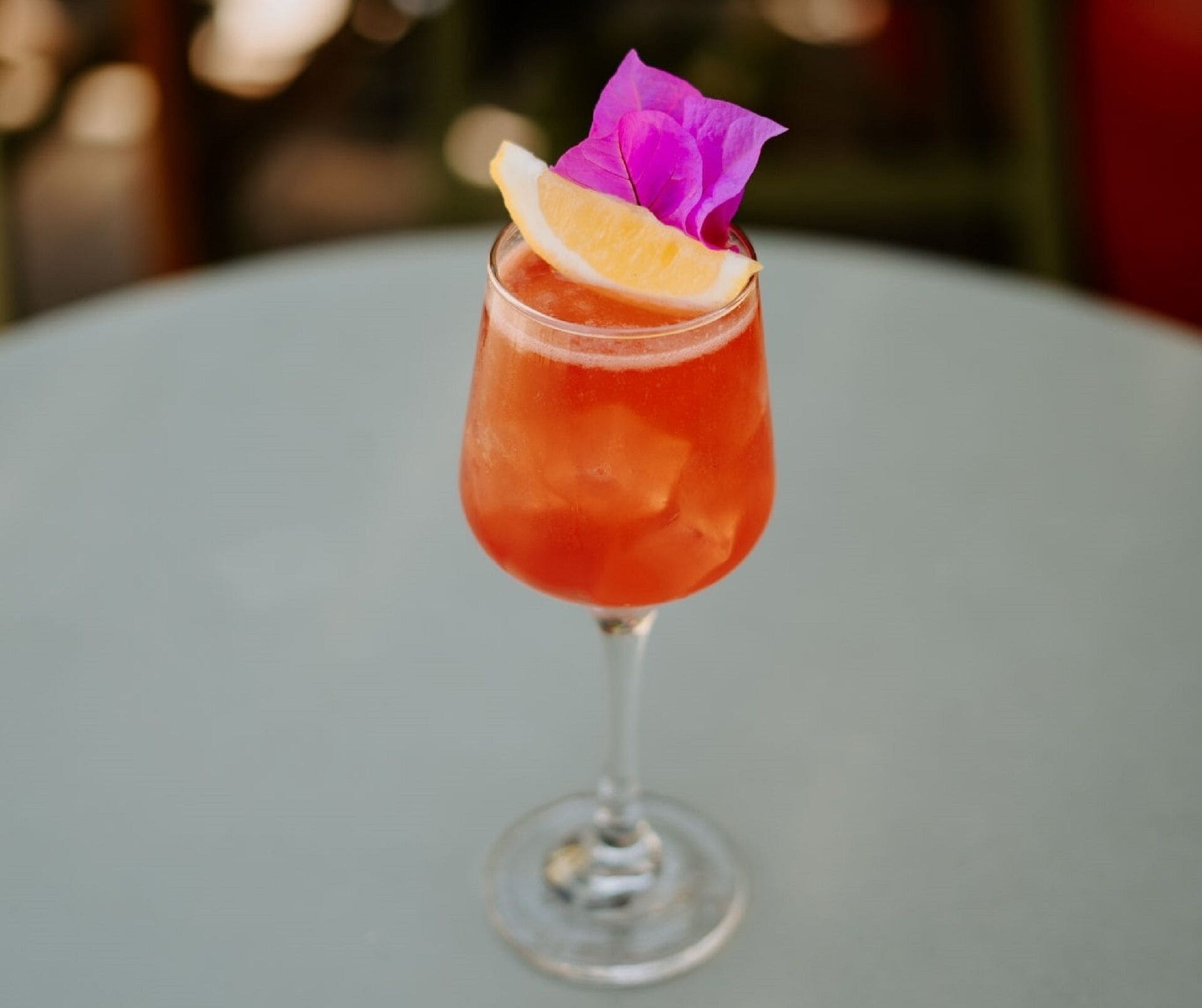 A cocktail in a stemmed glass with a slice of orange and a purple flower garnish on a table.