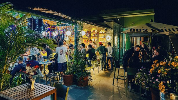 An outdoor dining area with tables and chairs, surrounded by potted plants and a bar area with people. - Viajero Oaxaca Hostel (Oaxaca)