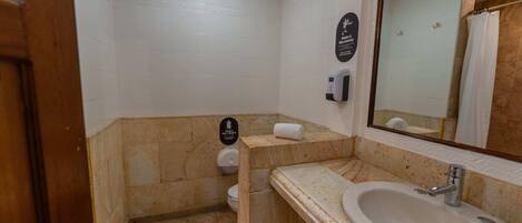 Un baño con lavamanos de mármol, inodoro, secamanos y espejo.
