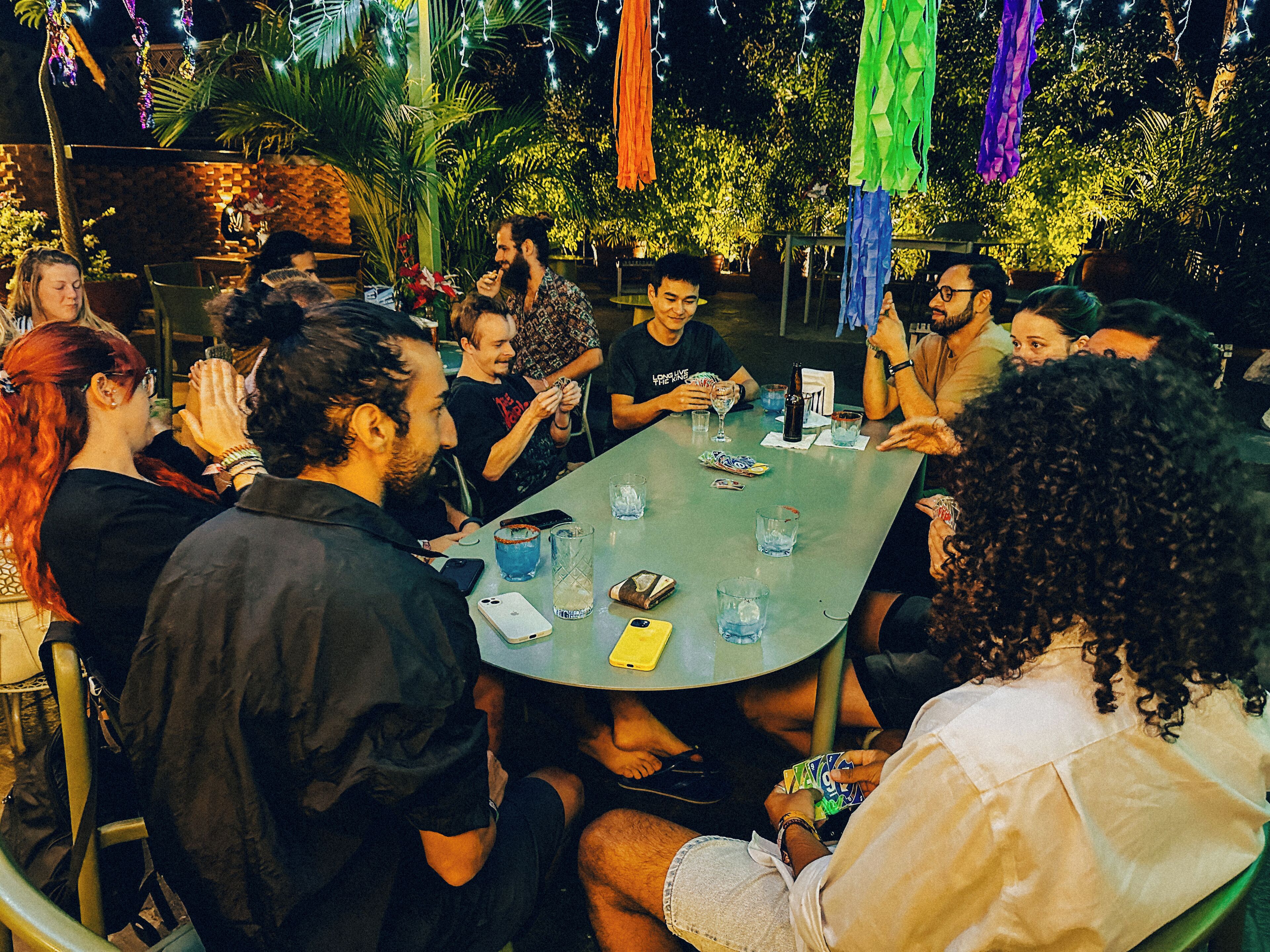 Un grupo de personas sentadas alrededor de una mesa, con bebidas y una botella, en un espacio exterior con plantas y luces colgantes.