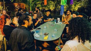 Un grupo de personas sentadas alrededor de una mesa, con bebidas y una botella, en un espacio exterior con plantas y luces colgantes.