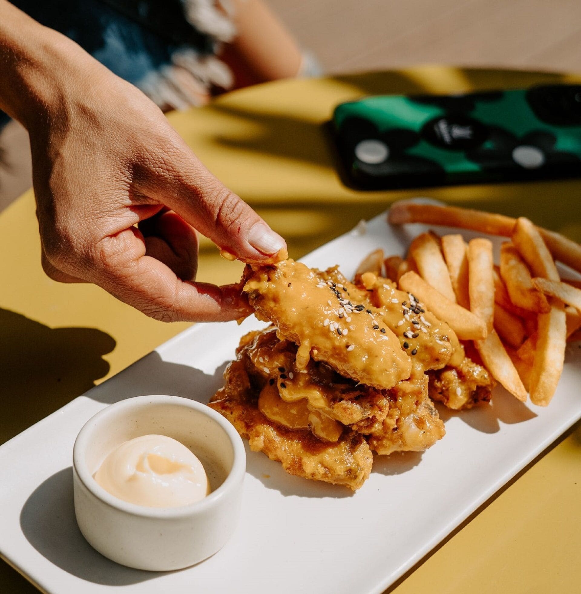 Una mano sosteniendo un pedazo de pollo frito acompañado de papas fritas y un recipiente pequeño con salsa.