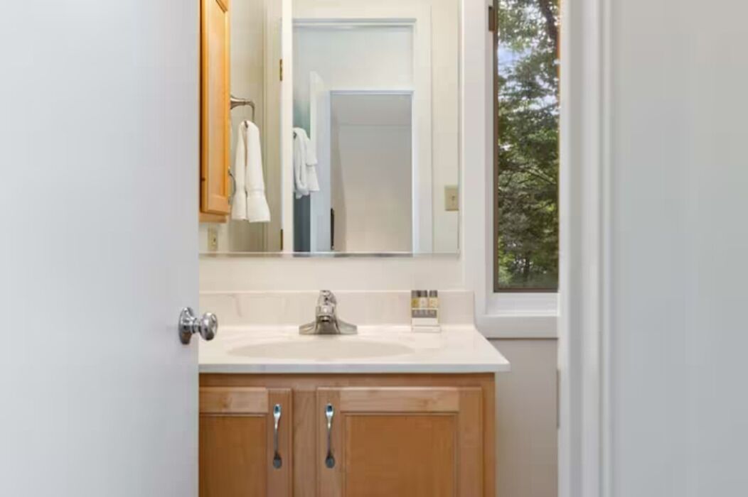 Un bagno con lavandino bianco, mobili in legno, uno specchio e una finestra vista alberi.