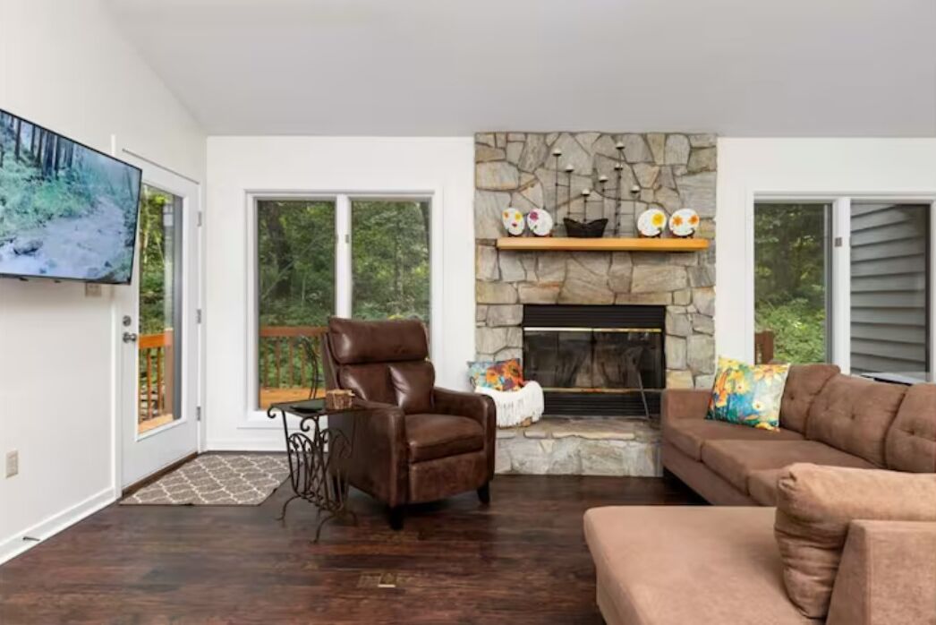 A living room with a stone fireplace, a brown leather chair, a brown sofa, and a large TV on the wall.