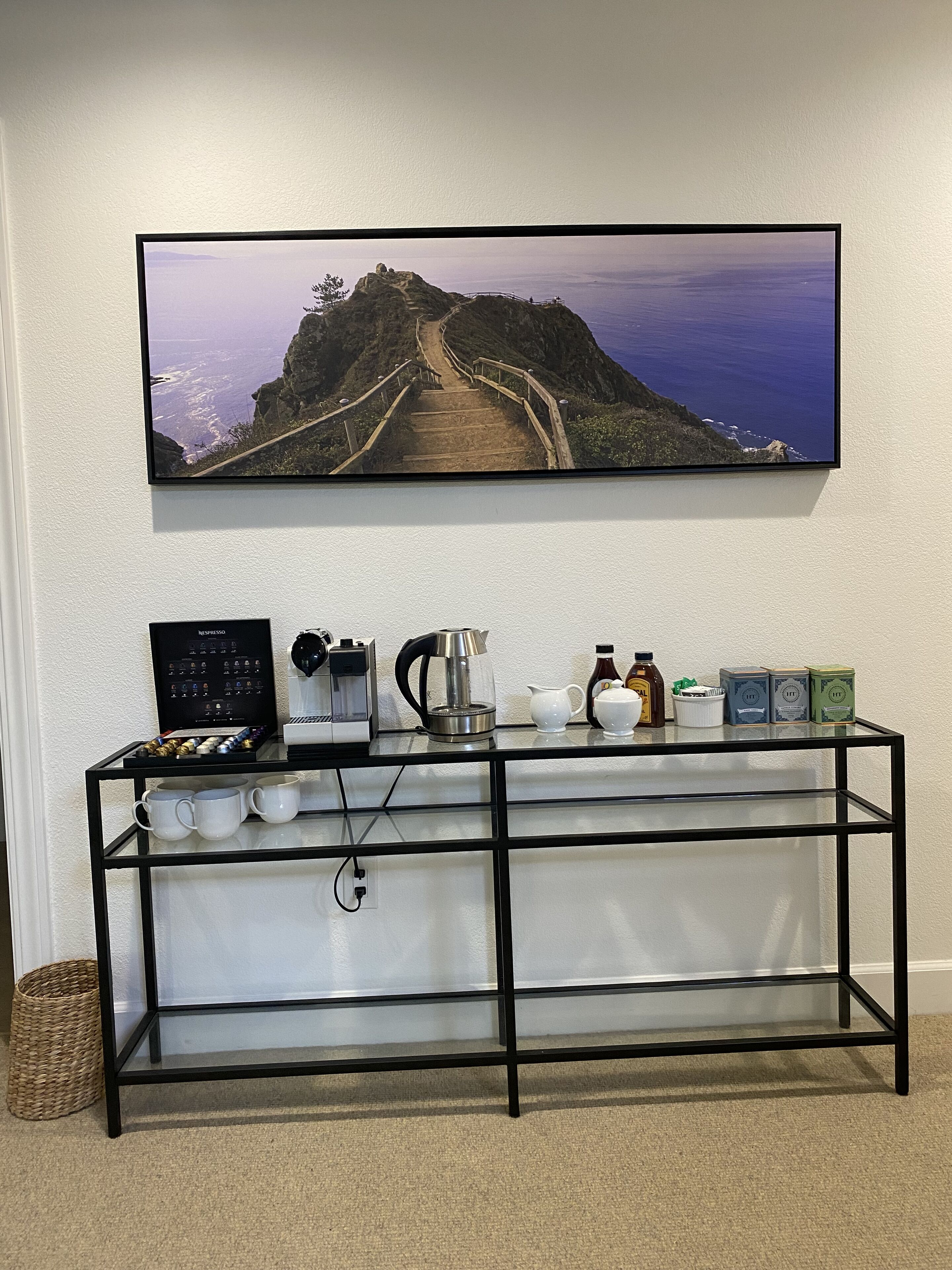 A coffee station with a coffee maker, cups, and condiments on a shelf, with a scenic view of a coastal path above.