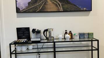 A coffee station with a coffee maker, cups, and condiments on a shelf, with a scenic view of a coastal path above.
