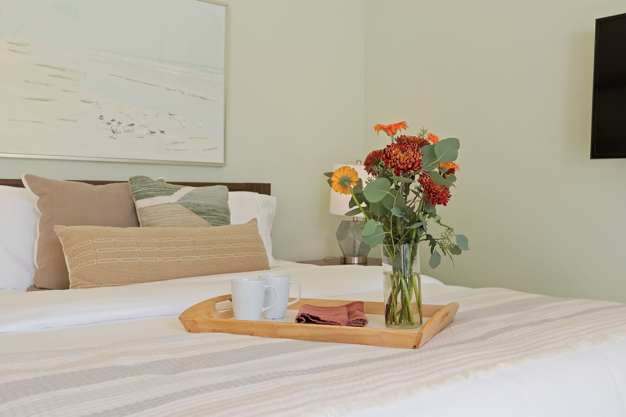 Ein ordentlich bezogenes Bett mit einem Tablett, das zwei weiße Tassen, ein zusammengerolltes Handtuch, eine Vase mit Blumen und ein Bild an der Wand zeigt.