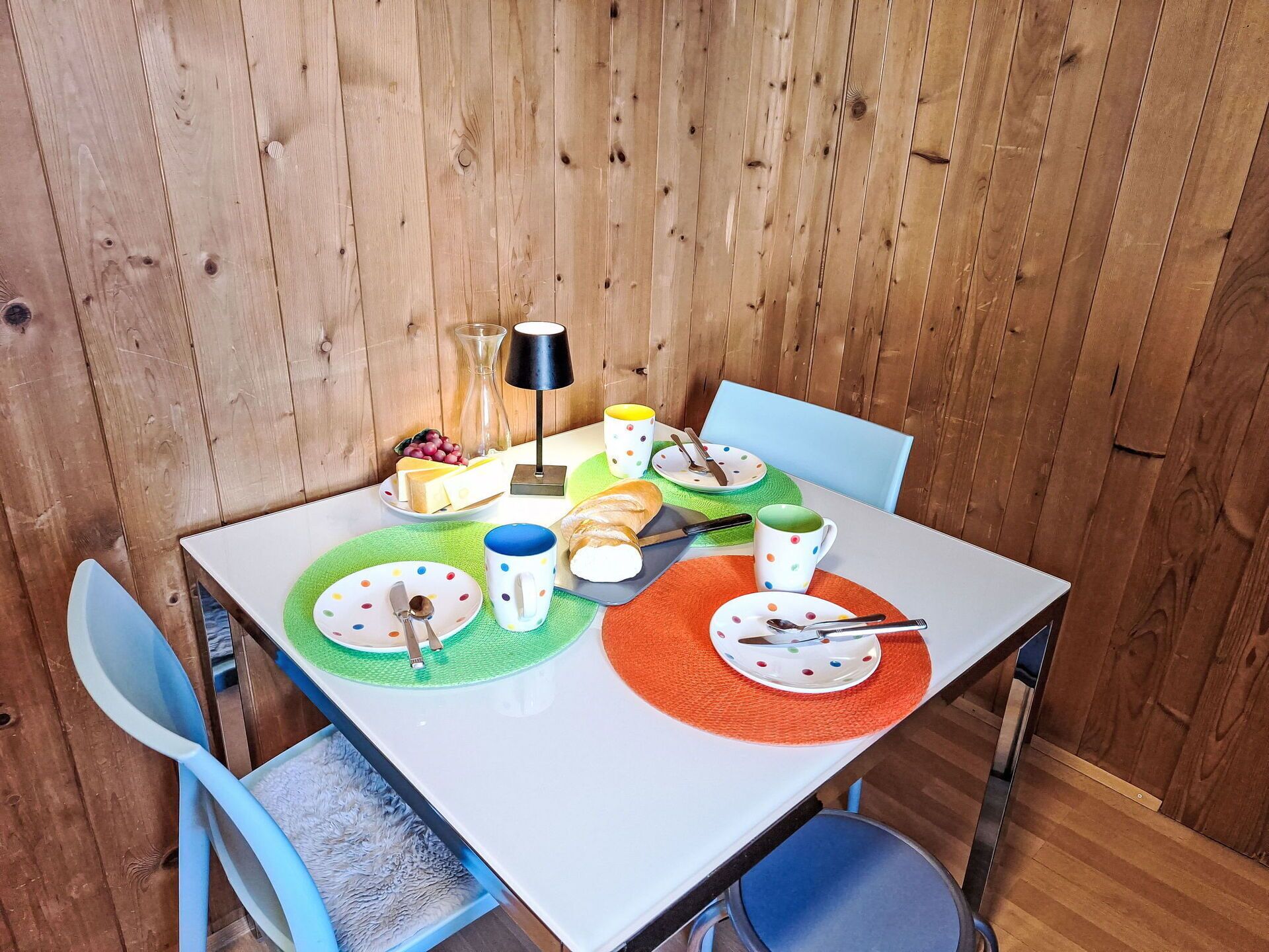 A dining table set with plates, cutlery, and a lamp, surrounded by blue chairs and a wooden wall.