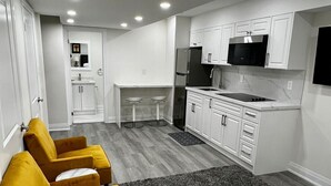 A modern hotel room with a bed, a seating area with yellow chairs, a kitchenette, and a small desk.