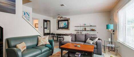 Una sala de estar con un sofá verde, una mesa de centro y un sillón amarillo. Se ve una escalera que sube a otro nivel, un comedor con una mesa y sillas, y un televisor montado en la pared.