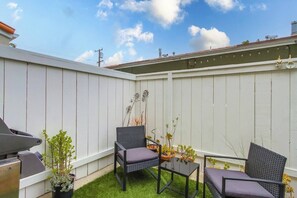 A backyard with a white fence, two wicker chairs, a small table, and a grill.