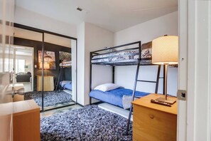 Een compacte hotelkamer met een stapelbed, een klein bureau met lamp en uitzicht op een zithoek door een glazen deur.