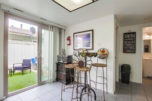 A room with a sliding glass door leading to a backyard, a dining area with metal chairs, a framed picture on the wall, and a sign with beach-related items.
