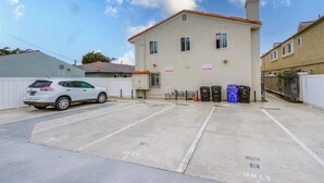 A parking lot with a white car parked in the foreground, a building with multiple windows and a red roof, and several trash bins lined up against the building. - Pet-friendly 1 block to the beach with parking (San Diego)