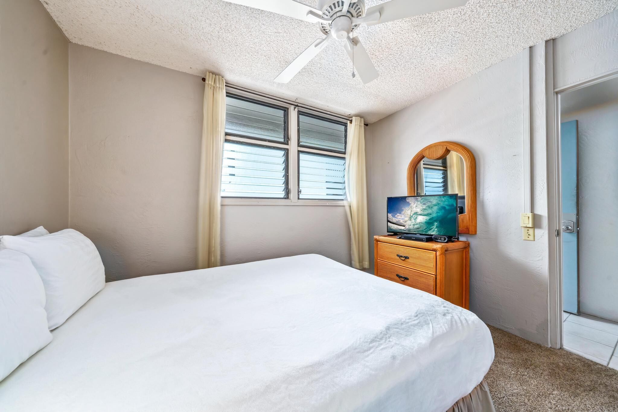A hotel room with a bed, a wooden dresser, a television, a ceiling fan, and a window with curtains.