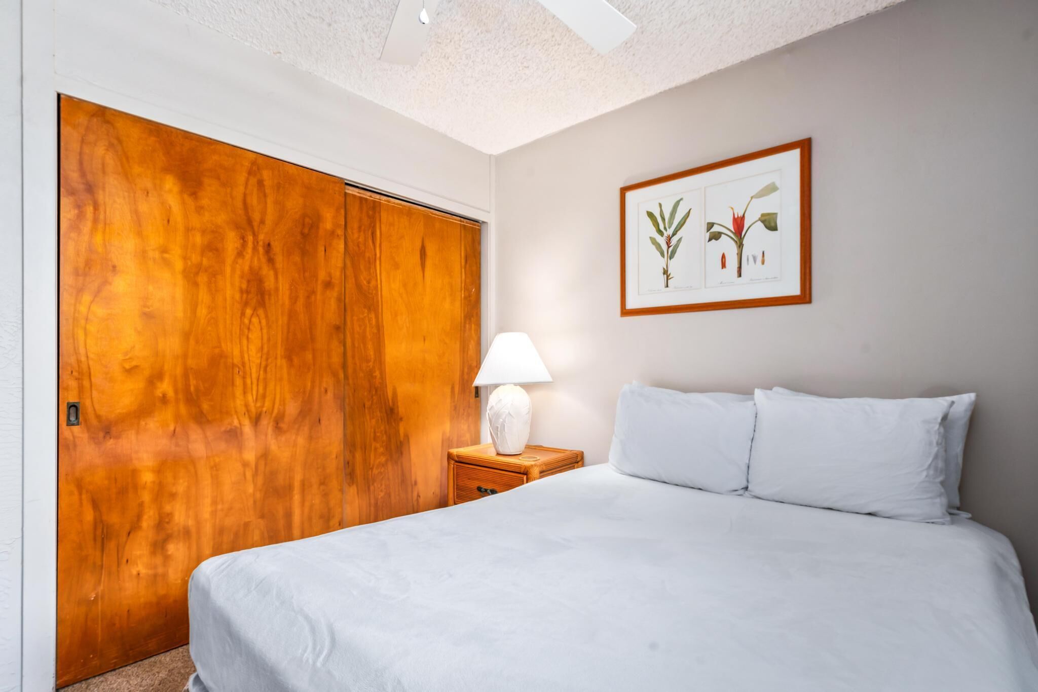 Un dormitorio con una cama, una mesita de noche con lámpara, un armario de madera e ilustración botánica enmarcada en la pared.