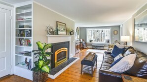 A living room with a fireplace, bookshelves, a leather sofa, and a large window. - Quiet Waterfront Home: Ideal Maine Escape! (Bass Harbor)