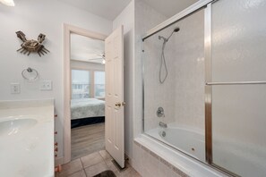 A bathroom with a bathtub, shower, and a door leading to a bedroom with a bed and windows.