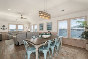 A spacious living area with a large sofa, a dining table with chairs, and a view of the water through large windows.