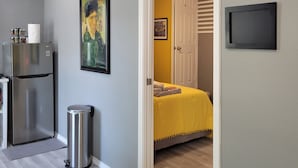 Un espacio habitable compacto con área de cocina, una cama con colcha amarilla, una puerta que da a un baño y un televisor montado.