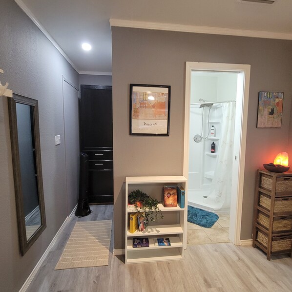 Un pasillo con una puerta que da a un baño, un cuadro en la pared, un estante pequeño con libros y un objeto decorativo sobre una mesa.