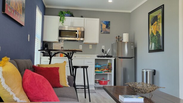 Una cocina moderna con armarios blancos, un refrigerador de acero inoxidable y un microondas. Hay un comedor con una mesa y sillas, y una sala de estar con sofá y cojines.