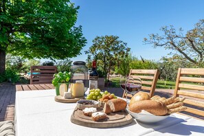 Un montaje para comer al aire libre con una mesa, sillas de madera, una botella de vino, queso, uvas, pan y un bol de aceitunas.