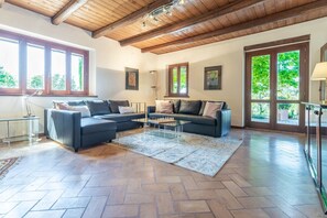A spacious living room with a wooden ceiling, two sofas, a coffee table, and large windows offering a view of greenery.