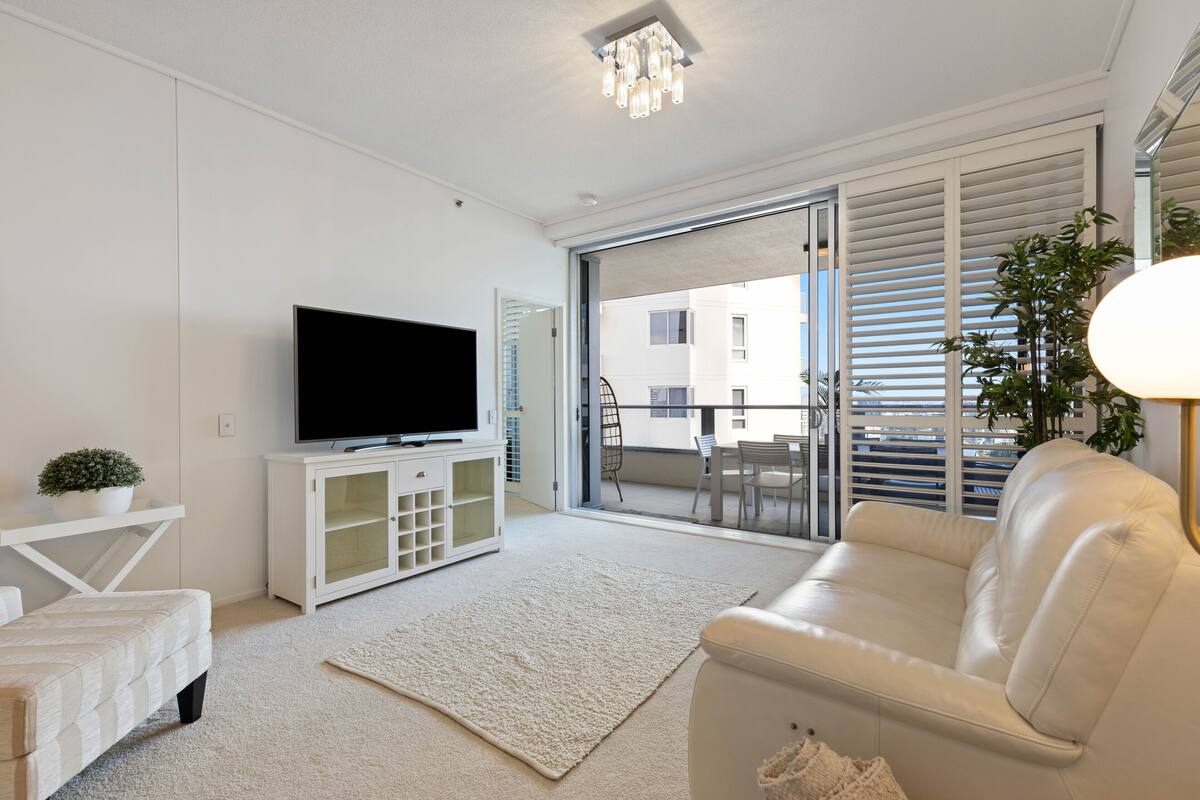 A modern living room with a flat-screen TV, a white sofa, a small table with a plant, and a sliding glass door leading to an outdoor balcony.