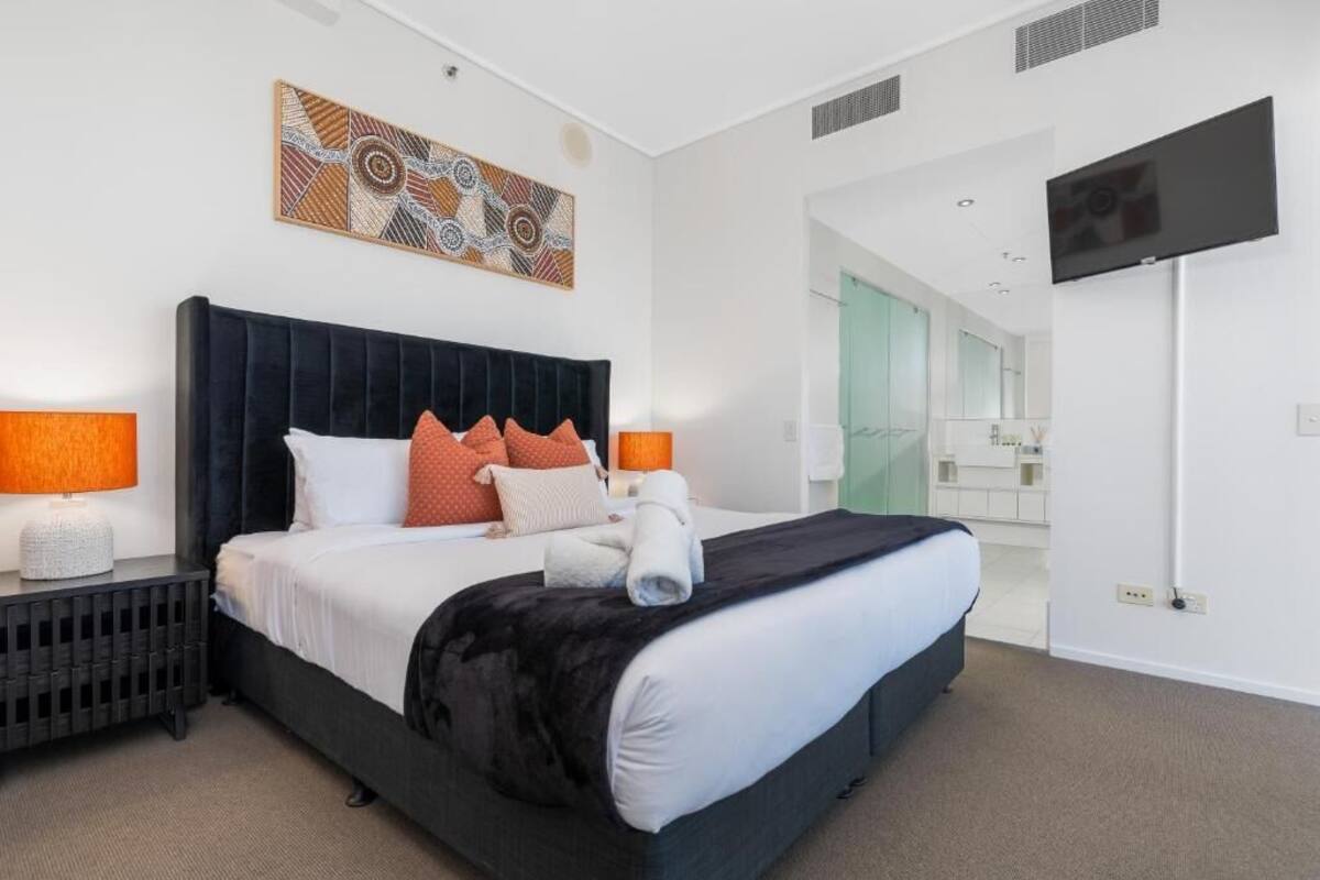 A modern hotel room with a large bed, two bedside lamps, a flat-screen TV, and a bathroom visible through an open door.