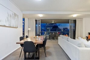 Una habitación de hotel moderna con comedor, sofá y vistas a un paisaje urbano al anochecer.