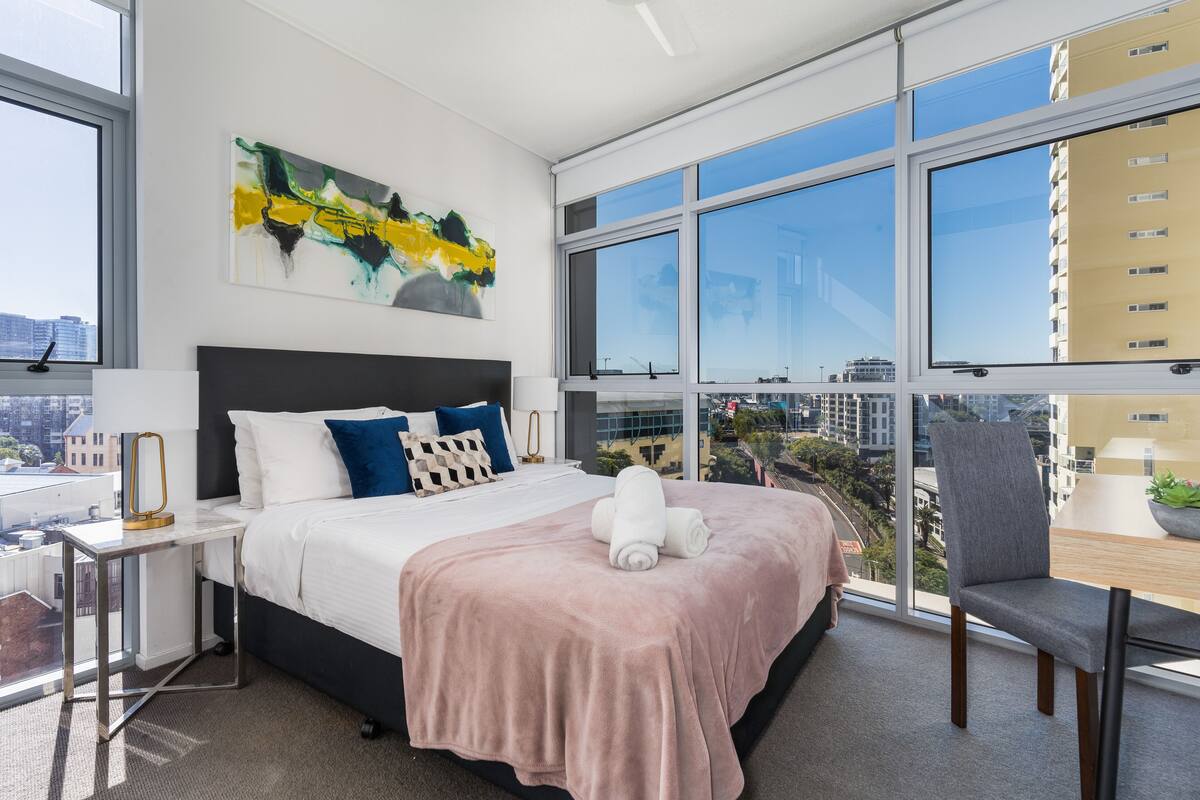 A modern hotel room with a large bed, a desk, and a chair. There is a large window with a view of the city.