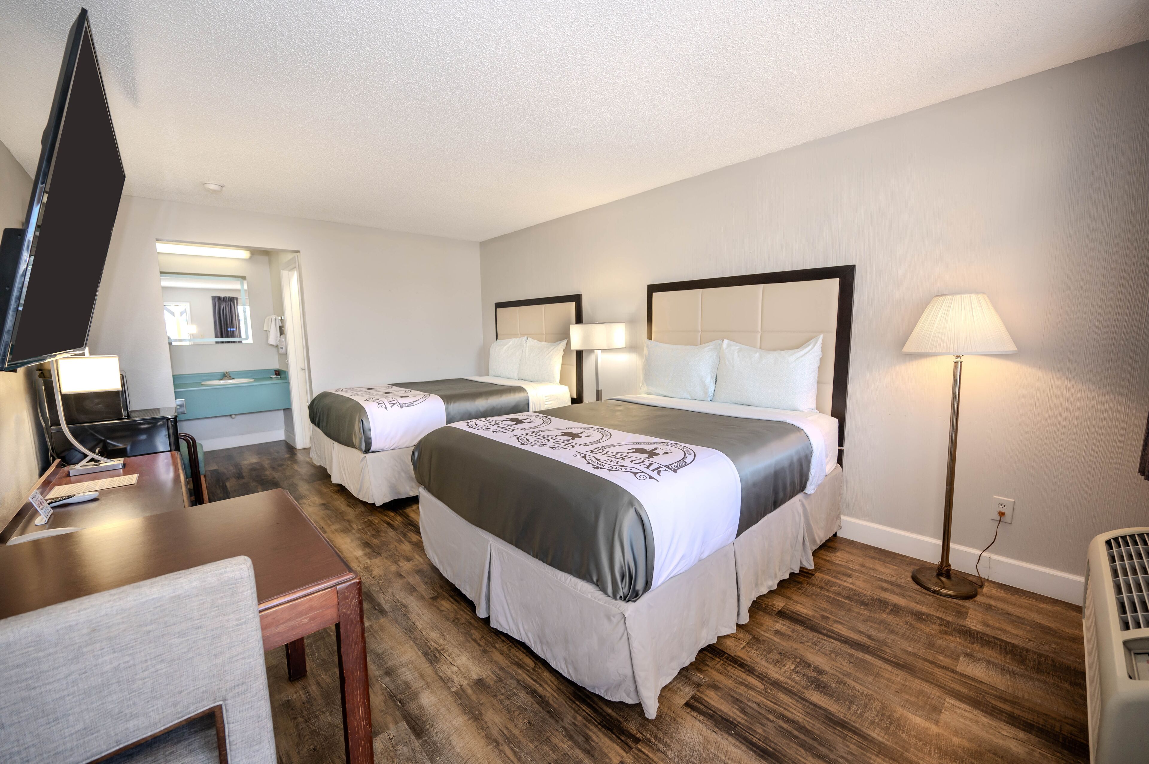 A hotel room with two beds, a desk, a chair, a TV, and a bathroom visible through an open door.