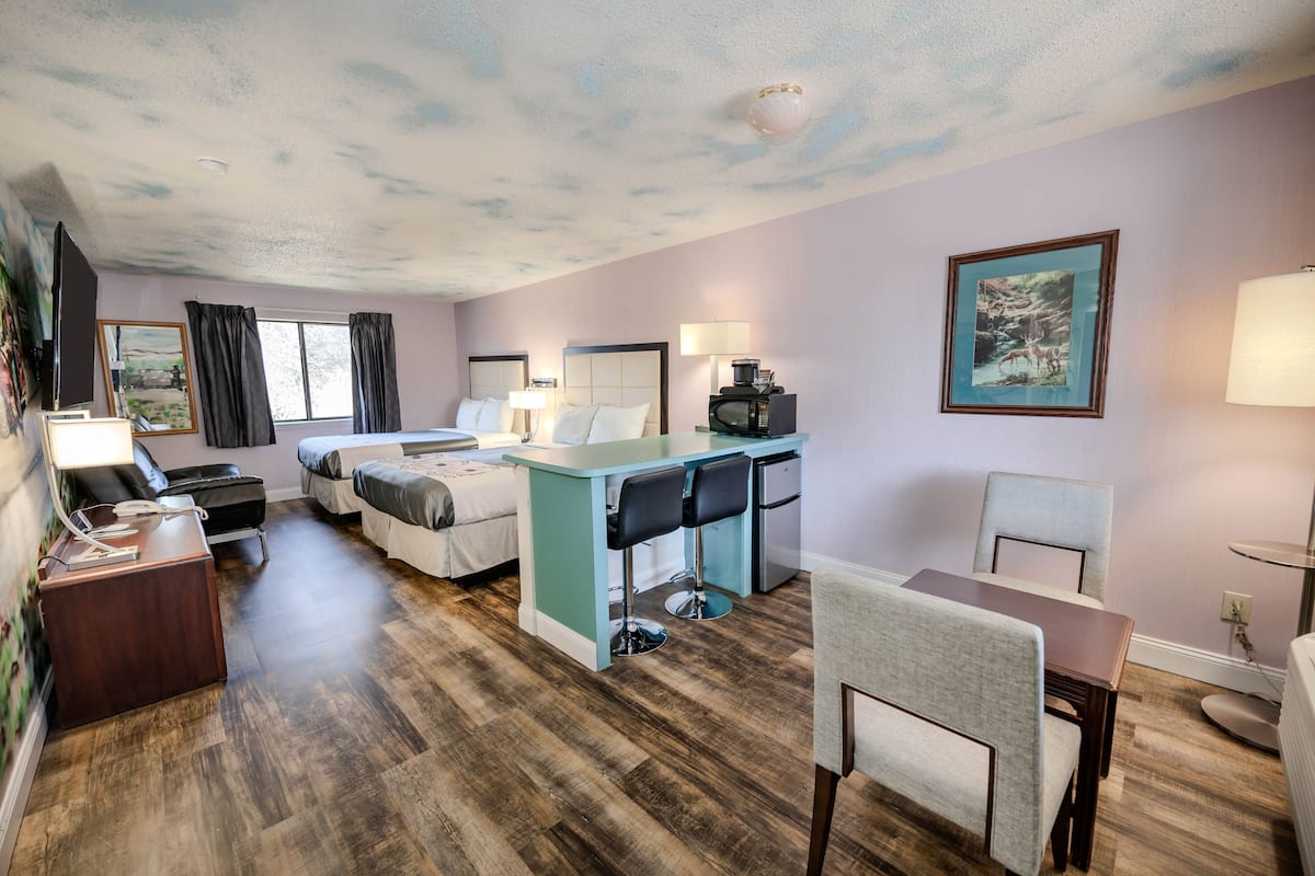 A hotel room with a bed, a desk with two chairs, a small table, a TV, and a painting on the wall.