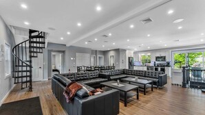 Ein geräumiger Wohnbereich mit einem großen, L-förmigen Ecksofa, einem schwarzen Couchtisch und einem schwarzen Akzentstuhl. Im Hintergrund ist eine schwarze Wendeltreppe zu sehen.