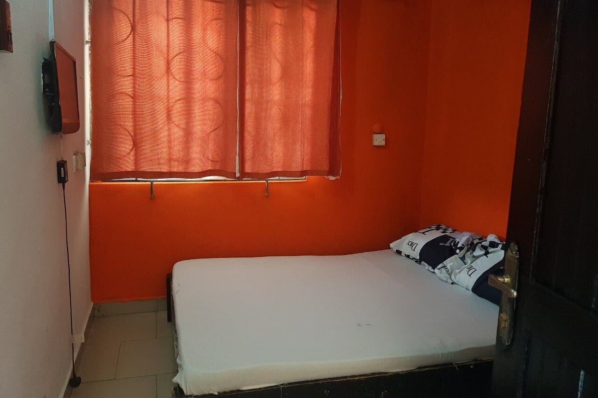 A small room with an orange wall, a single bed, and a window with curtains.