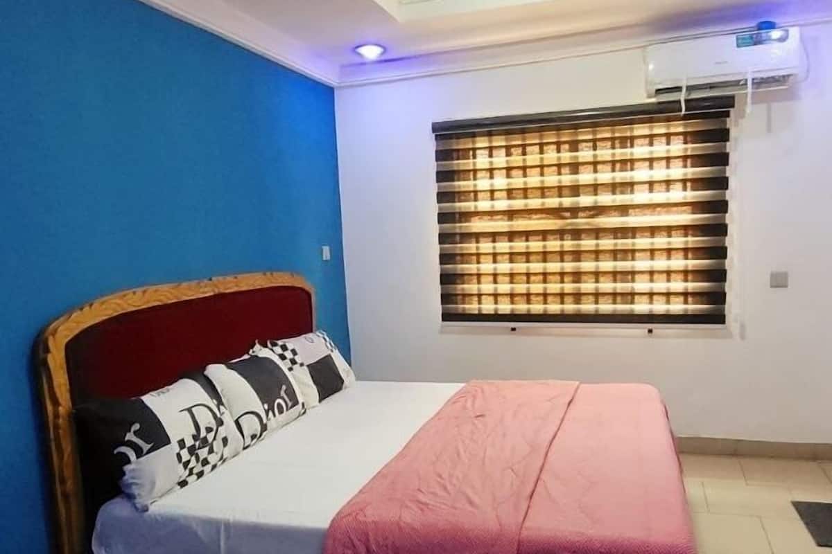 A bedroom with a bed, a window with blinds, an air conditioning unit, and a blue wall.