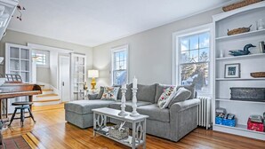 A living room with a piano, grey sofa, white side table, and a bookshelf with decorative items. - Charming beach home, complete with pool & cozy guest house! (Clinton)