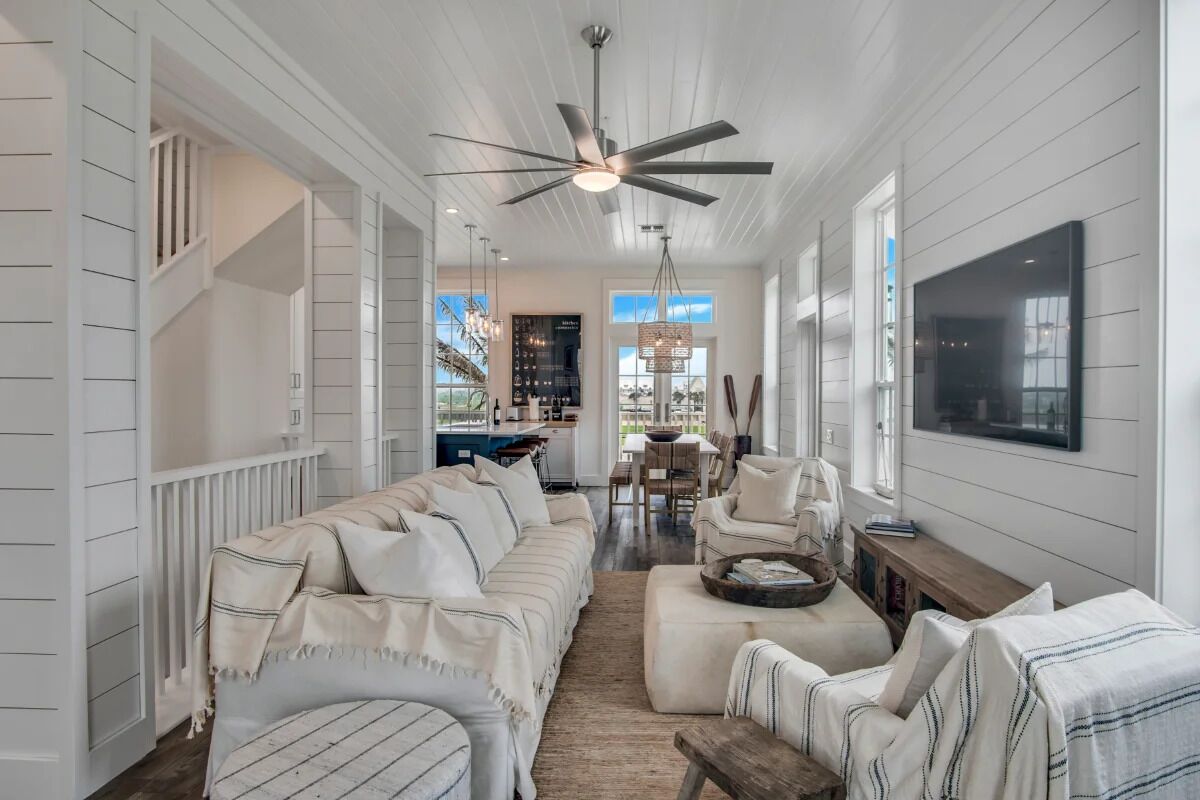 A spacious hallway with a high ceiling fan, white wooden walls, a large sofa, a dining area, and a kitchen visible through an open door.