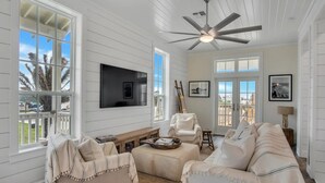 A spacious living room with a ceiling fan, large windows, a flat-screen TV, and a view of the outdoors. - Shared Heated Pool, Golf Cart Boardwalk to Beach (Port Aransas)