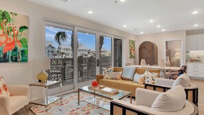 Un salón espacioso con un gran ventanal que da a un balcón, un sofá beige, una mesa de centro y una zona de comedor con alfombra estampada.