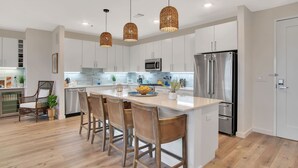 Una cocina moderna con armarios blancos, una isla central con taburetes de bar, electrodomésticos de acero inoxidable y lámparas colgantes de mimbre.