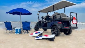 Eine Strandlandschaft mit einem Golfwagen, zwei blauen Liegestühlen, einer blauen Kühlbox und einem weißen Brettspiel.