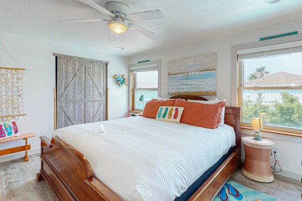 A bedroom with a wooden bed, a ceiling fan, a window with a view, and a wall-mounted shelf with decorative items.