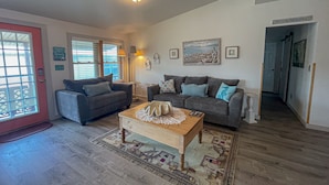 Ein Wohnzimmer mit zwei Sofas, einem Couchtisch und einem Teppich. An der Wand hängen gerahmte Bilder und eine Tür mit einem Schild.
