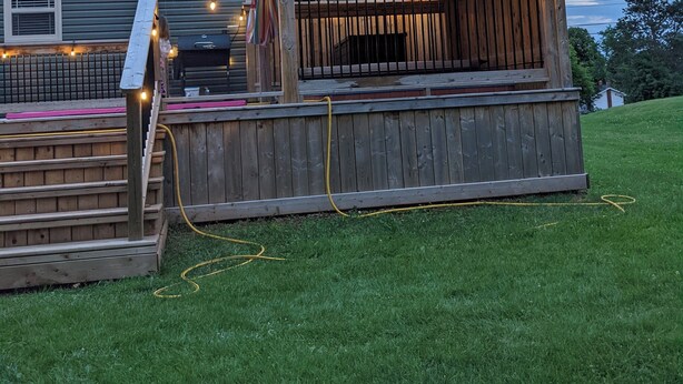 Eine überdachte Terrasse im Freien mit Lichterketten, Holelllaufzaun und einem Gartenschlauch auf einer Rasenfläche.